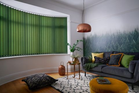 Reber forest vertical blinds in a bay window. In a living room with wallpaper on the wall and a dark sofa to the right side and a coffee table next to the sofa. 