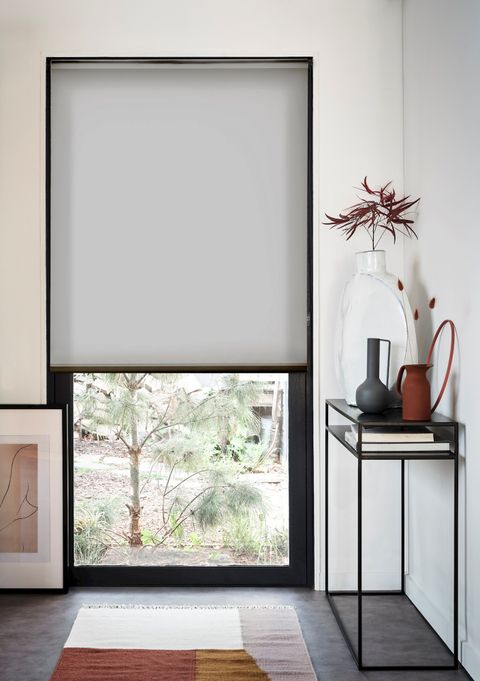 reber ash roller blinds at a tall window in a white room and a tall black table on the right with vases on top 