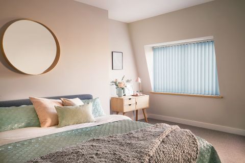 Reber Powder Blue vertical blinds in a cream bedroom. A bed on the left hand side with a mirror above it and a desk in the corner of the room. 