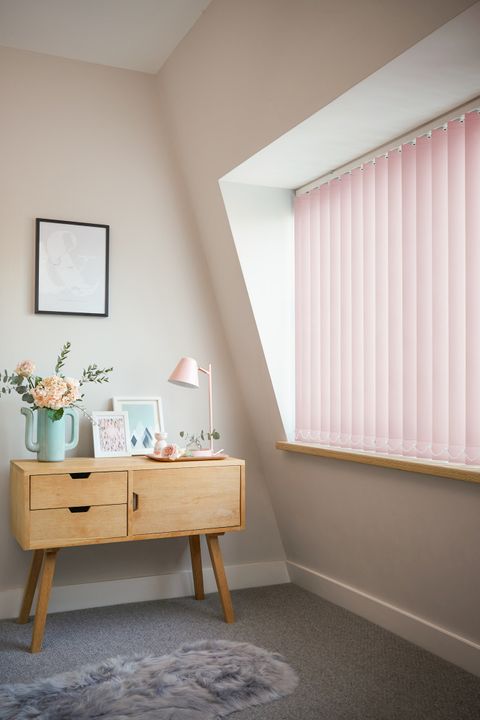 Reber blush vertical blinds in a bedroom with a desk and drawers next to the window. Flowers, framed pictures and a pink lamp are on the desk. 