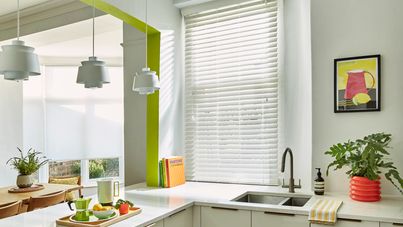 Mirage White Maple blind in a white kitchen with lime features. There is an attached breakfast bar with a tray of breakfast food and drinks on it. There is a sink with a plant next to it in the right side and a wooden table with chairs on the left side.