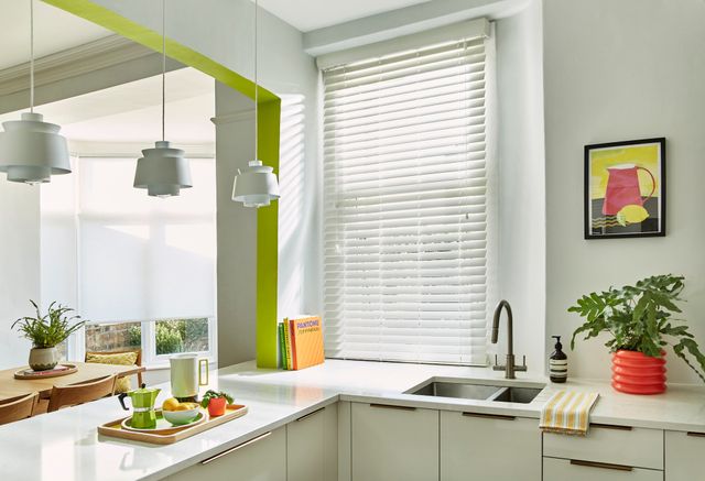 Mirage White Maple blind in a white kitchen with lime features. There is an attached breakfast bar with a tray of breakfast food and drinks on it. There is a sink with a plant next to it in the right side and a wooden table with chairs on the left side.