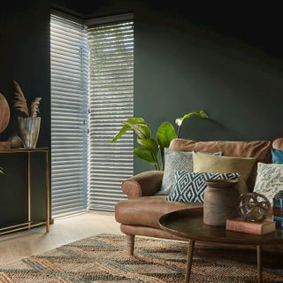 wood venetians fitted to two tall windows in a living room decorated with dark green walls and a brown leather sofa