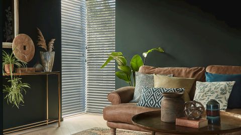 wood venetians fitted to two tall windows in a living room decorated with dark green walls and a brown leather sofa