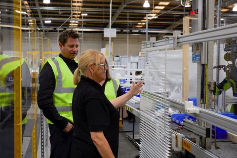 Woman looking at white venetian blind in a factory while two people in hi-vis watch