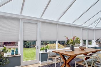 crush silver grey conservatory pleated blinds by a wooden dining table and numerous plants