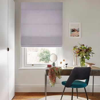 Faso Lavender Purple roman blind on a window in a white decorated room that has a dressing table, chair & wardrobe.
