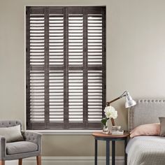 black ash coloured shutters in a bedroom inbetween a grey chair and bed on an off white wall