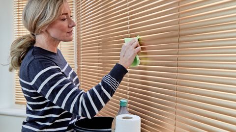 lady with blonde hair using sponge cloth bucket and paper towels to clean metal venetian blind