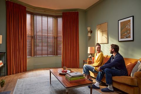 orange curtains paired with brown faux wooden blinds on large bay window in cosy living room with two people chatting on the sofa