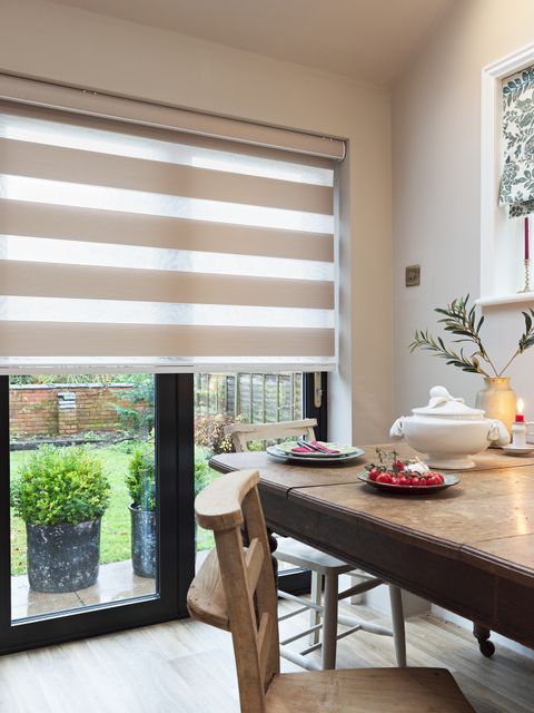 Cream dining room with small window dressed in Roman blind in white with green foliage pattern and main windows and door to rear with beige day and night blind, lowered halfway, rustic wooden dining table and chairs, red candles.