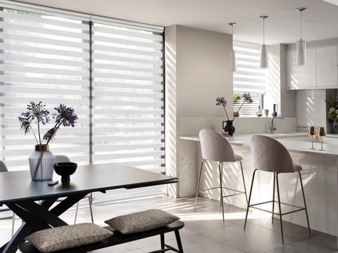Kitchen/diner in white with floor to ceiling windows and doors in dining are and window in kitchen area all dressed in white day and night sheer blinds, fully lowered in dining area, kitchen halfway lowered, dark wood table and bench with cushions, white units with bar stool chairs, grey tiled floor with rays of light.
