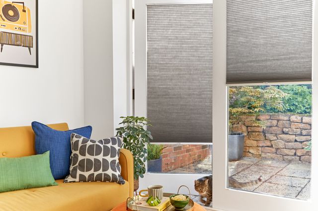 White living room with French doors dressed in dark grey duette blinds, left hand three quarters lowered and door open, right hand lowered halfway and door closed, a cat slinks in the gap to enter the room with a saffron yellow sofa with a green cushion, blue cushion and cream cushion with dark grey semic circular pattern, orange poufee with cup and book.