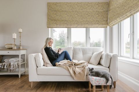 Corner of ivory living room with windows to both walls into corner, dressed in Roman blinds with a wheat-coloured frond pattern on a pale beige background, lowered partially, a woman reclines on a cream two-seater sofa, looking at a grey cat lying on a footstool by the sofa, a white painted side dresser with two wooden candlesticks with lit pillar candles and a folded throw on a shelf below, mid wooden floor.,