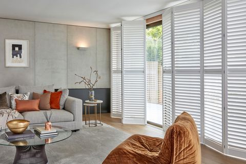Grey living room with wooden floor and grey rug, grey sofa with cushions in browns and coppers, glass circular coffee table, copper soft chair and floor to ceiling windows and doors in white tracked shutters, partially open.