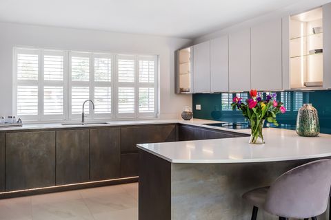 White kitchen with dark brown units to three walls, white shutters behind sink, closed with louvres open, white worktops extending to L-shaped seating area with additional units underneath, white wall units to right above cobalt blue splashback.