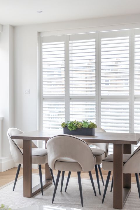 Dining area with white vinyl shutter on patio doors, closed, louvres open, wooden table and modern chairs in front, window with raised white Roman blind to left, contemporary grey sofa in left foreground.