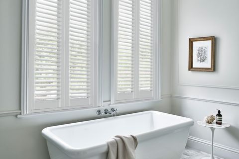 Bathroom in white with, tall window dressed in full height white vinyl shutters, closed with louvres open, bath beneath with taps on wall, small round white pedestal table with toiletries, beige towel over bath.