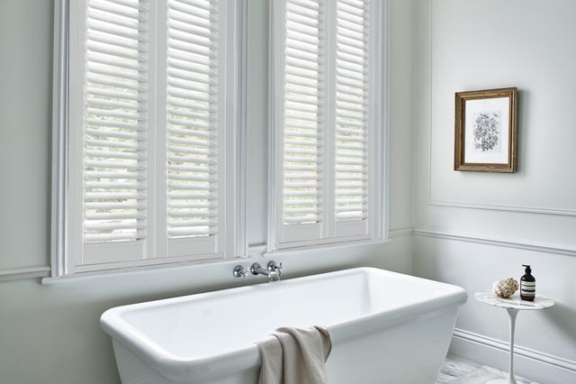 Bathroom in white with, tall window dressed in full height white vinyl shutters, closed with louvres open, bath beneath with taps on wall, small round white pedestal table with toiletries, beige towel over bath.