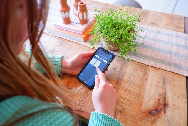 Close up on a woman sitting at rustic wooden table with a striped runner in pale blue and beige, small plant in terracotta pot, the woman is using an app on her phone to control roller blinds out of sight.