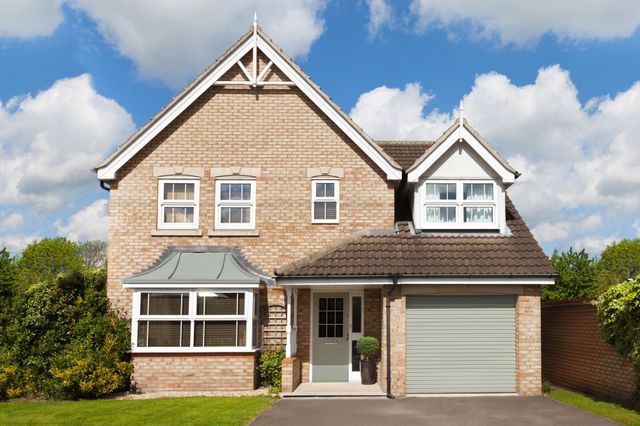 Exterior shot of large modern brown-brick house with integral garage door in pale grey-green, closed, white fascias to box bay window on ground floor, three small windows above bay and double window above garage, asphalt driveway with lawn to left and hedging.