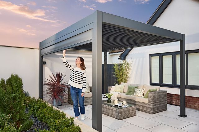 Exterior in a garden of a freestanding pergola in grey at dusk, installed on a cream patio next to a cream rendered house and a raised flower border with alpine plants and a raffia style sofa and coffee table under the pergola, a woman pulls down the side panel on the left.
