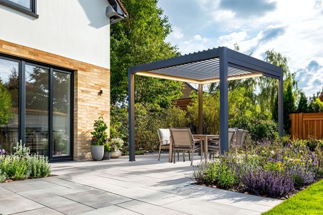Exterior of a modern house, with a large paved patio down to a lawn with small inset flower beds at random points in the patio and a freestanding grey pergola with louvred roof over four chairs and a table,