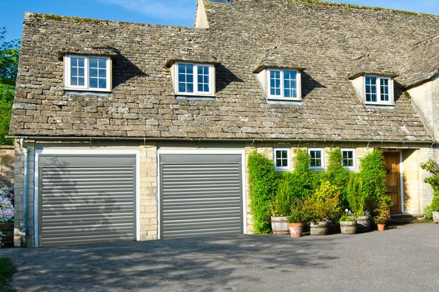 Large traditional pale stone house with wide driveway and integral double garage on left with two grey garage doors.