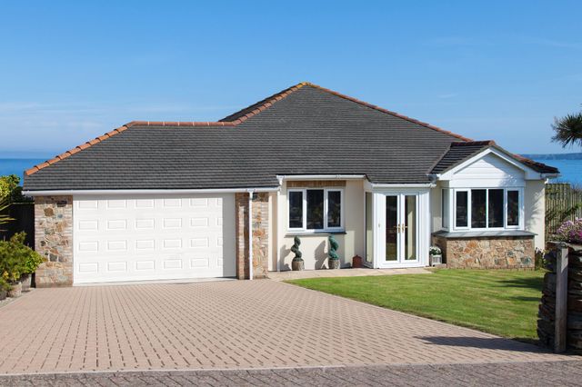 Exterior shot of modern brown-brick bungalow with integral garage door with georgian-effect panelling in white, closed, matching front door, white fascias to windows, brick driveway with lawn to right and flower pots to left of drive.