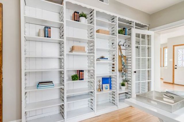 A home office with wooden floor, floor to ceiling white shelving with a mix of empty shelves and books, stationary and plants.