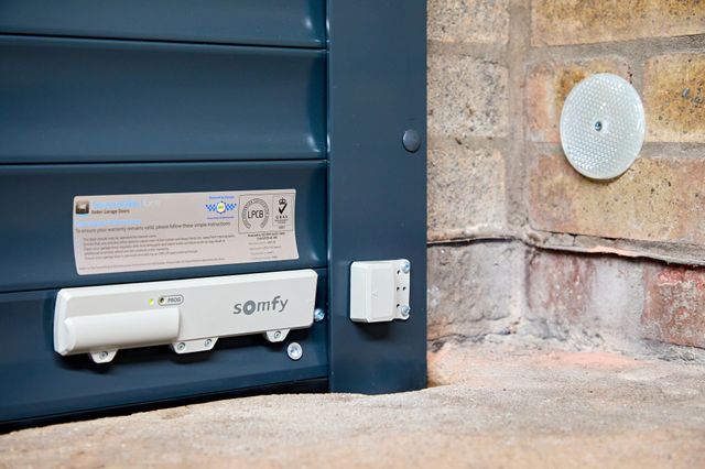 Close up of the bottom of a grey garage door, showing a somfy motor, locking system and a secured by design plaque.