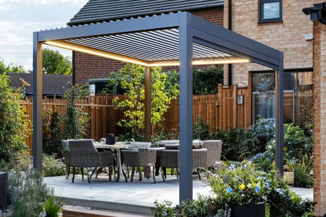 Exterior of a modern house, with a large paved patio down to a lawn and a freestanding grey pergola with fabric roof over four chairs and a table on a raised section of patio, with two grey rattan sofas at 90 degree angle with cream cushions.