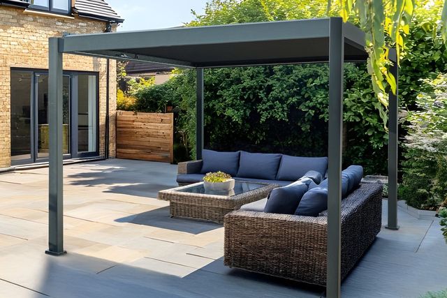 Exterior of a modern house, with a large paved patio down to a lawn and a freestanding grey pergola with fabric roof over four chairs and a table on a raised section of patio, with two grey rattan sofas at 90 degree angle with cream cushions.