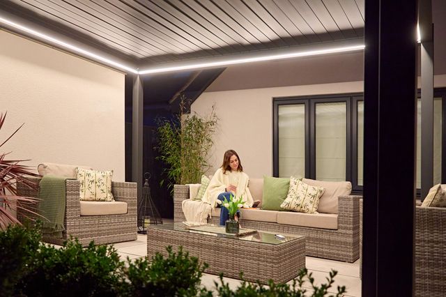 Close in on a woman sitting under an aluminium pergola with the louvres closed, at dusk with the lights on, on a rattan grey-brown sofa with matching chair and table.