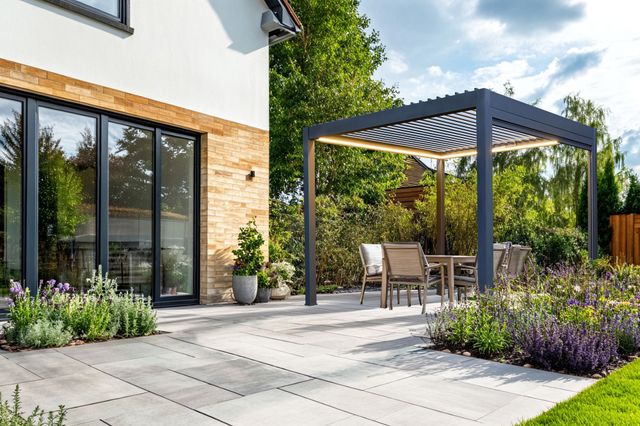 Exterior of a modern house, with a large paved patio down to a lawn with small inset flower beds at random points in the patio and a freestanding grey pergola with louvred roof over four chairs and a table,