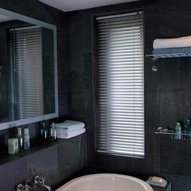 Silver venetian blind attached to a tall rectangular window in a bathroom decorated with dark slate tiles 
