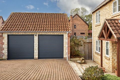Traditional yellow-brown brick house with adjacent double garage at 45 degree angle with anthracite roller garage doors, closed, a brick driveway connects the two, with a pergola porch over the house door which is a matching anthracite with four small square windows.