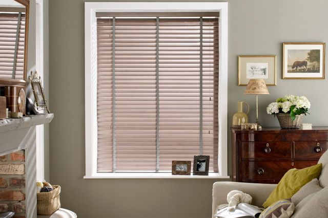 Living room in dove grey with window dressed in wooden venetian blinds in a mid tone with grey tape, beige sofa opposite stove in brick hearth, mide beige carpet, wicker log basket, mirror over white mantelpiece.