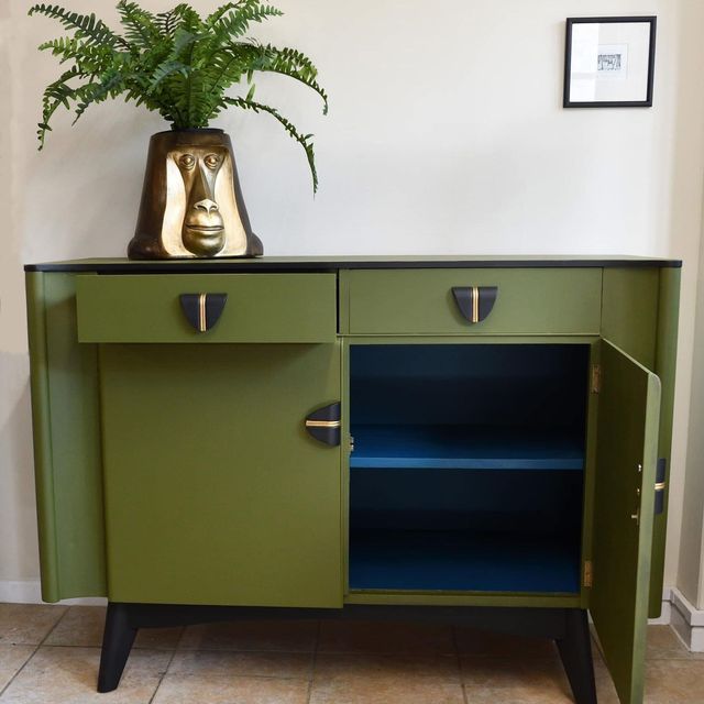 An olive green wood painted sideboard, right door open revealing cobalt blue painted interior, two drawers above, left partially open, all with black handles with gold stripe, plant pot in shape of primate in wood and gold with a fern in it.