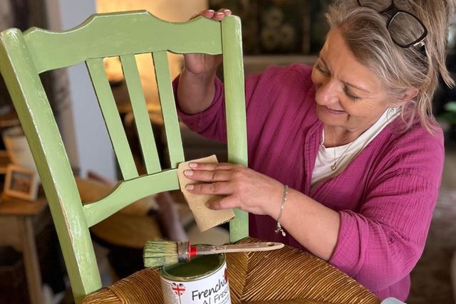 A woman wearing a dusky pink jumper sandpapers a wooden chair painted light green, an open tin of Frenchic paint and paintbrush on the seat.