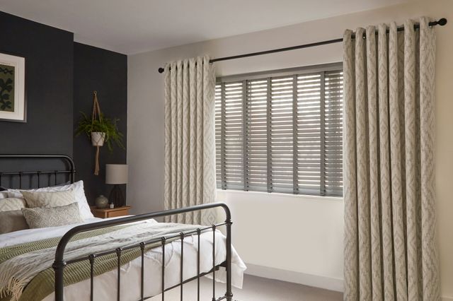 A modern bedroom with a minimalist, cozy design features a black metal bed frame with white and neutral-toned bedding, accented by textured cushions and a green throw. To the right, a large window is dressed with dark grey faux wood Venetian blinds and full-length curtains in a soft, patterned fabric. 