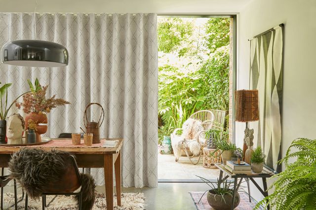 A cozy, eclectic dining area opens up to a lush garden through a large sliding glass door. On the left, a wooden dining table is adorned with natural decor including dried flowers, woven baskets, and pottery. A black chair with a dark fur throw sits at the table. Light grey wave-patterned curtains partially cover the glass door. On the right, a side table holds potted plants, books, and a sculptural lamp with a woven shade.
