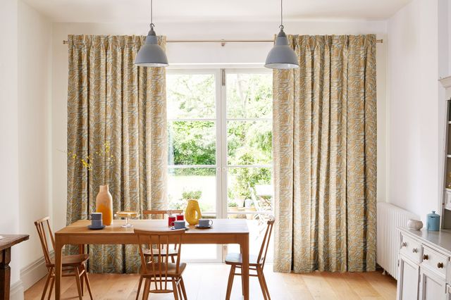 Dining room with cream walls, wooden floor, wooden dining table and chairs with grey cups and saucers, chartreuse jug and earthenware vase with yellow flowering twigs and French doors dressed in pinch pleat curtains with a chartreuse frond leaf pattern on a cream background.