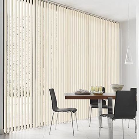White coloured vertical blinds are fitted to a wide and tall window in a dining room decorated in white and features a dining table and chairs