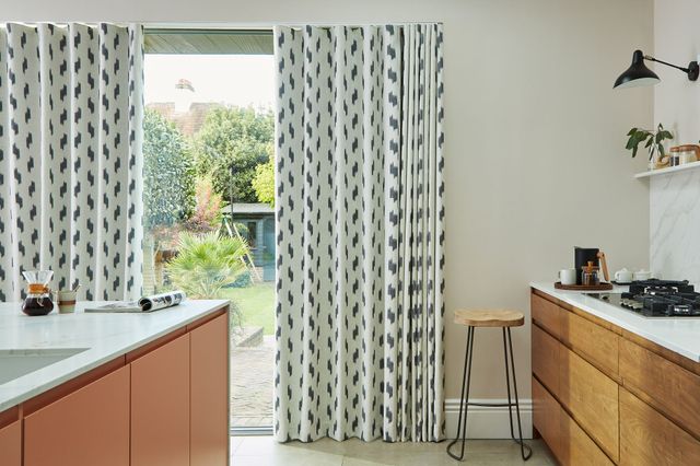 Modern kitchen with floor-to-ceiling glass doors partially covered by patterned curtains in a black and white geometric design. The kitchen features warm wooden cabinets, a white countertop, a gas stove, a minimalist bar stool with a wooden seat and metal legs, and a view of a garden outside.