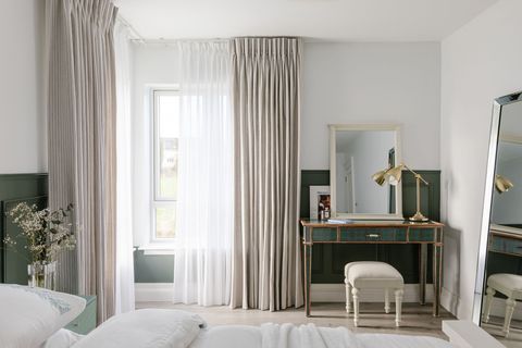 Chalk-white bedroom with two corner windows dressed in white voile curtains, partially pulled back, layered under pale oyster curtains, pulled back, bed in foreground with white bedding, dressing table and stool in corner against dark grey panelling. 