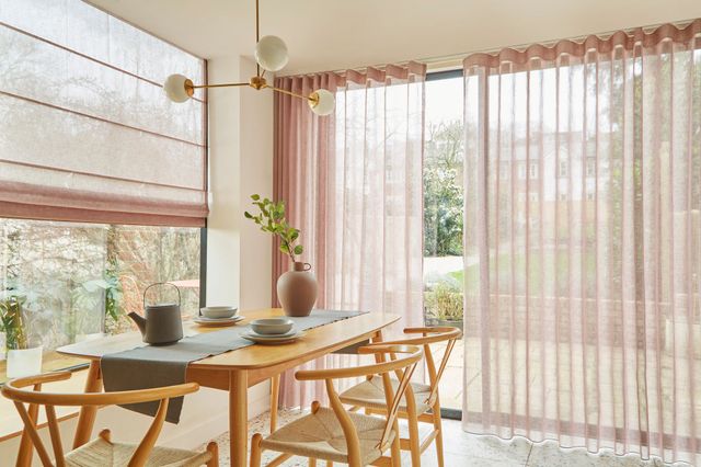Corner of cream dining room with picture window on left dressed in wide voile Roman blind in pale dusky pink, floor to ceiling window dressed in matching voile curtains, slightly parted, wooden modern table and chairs, grey runner, grey teapot, earthenware vase with foliage, tiled floor, pendant light with three arms and white spheres at end of each arm. 