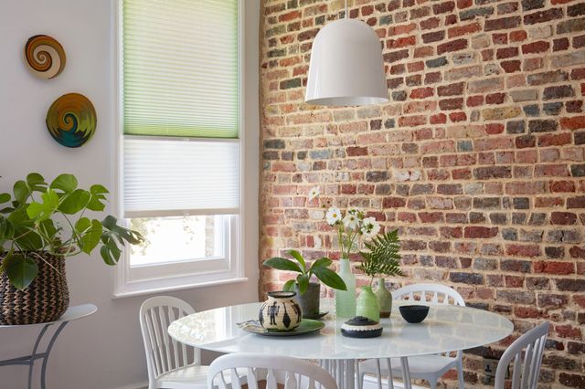 Corner of dining room with white wall on left with window dressed in two pleated blinds, pale green to top half and white blind cafe style to lower half, raised halfway, natural brick wall to right, round white modern dining table with white chairs, multiple vases and bowls in shades of green with a fern, ow-eye daisies and plant, console table with large pot with plant. 