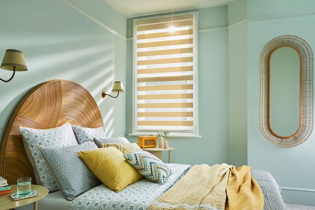 Bedroom with narrow tall window dressed in pale gold day and night blinds fully lowered with vanes tilted, bed with semi-circular wooden headboard, bedding in pale blue with dark blue geometric pattern, patterned cushions in blue and pale gold, pale gold throws, long oval mirror beside window. 