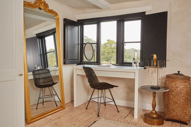 Room with wide shallow window dressed in black solid shutters, open, with black frames, floor to ceiling wide gilt-framed mirror leaning on wall to left, white desk and black chair at window, bronzed pedestal table to right, with necklace stand topped with a bird, high woven basket, black mirror on desk, pale wood floor with natural rug. 